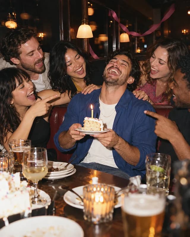 A man turning thirty laughs with his head back in a bar booth while his friends sing and point at him (AI photo, fictional people)