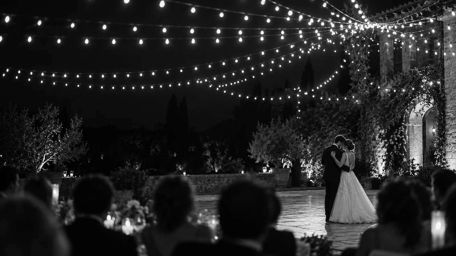 Black-and-white photo of a bride and groom dancing alone under hundreds of string lights in a courtyard at night (AI photo, fictional people)