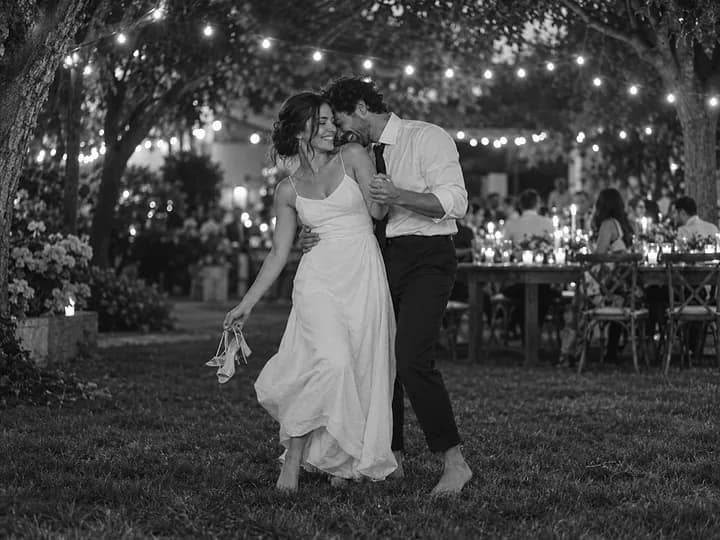 Black-and-white photo of a barefoot couple dancing on the grass in a garden strung with fairy lights, her shoes in her hand (AI photo, fictional people)