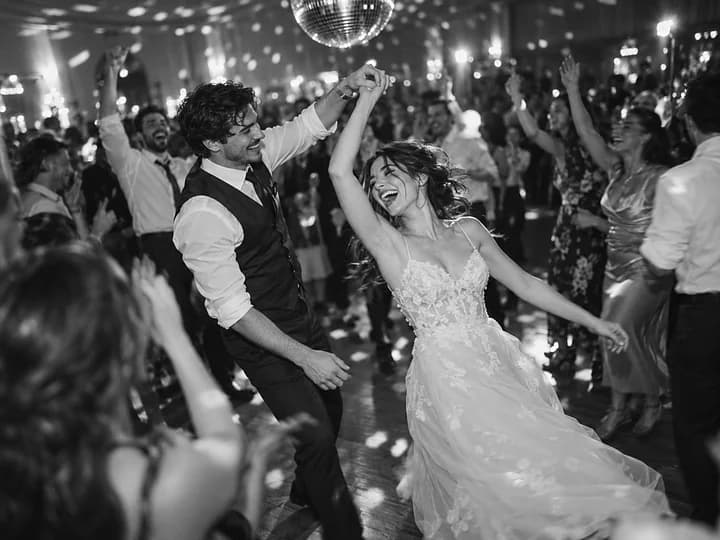 Black-and-white photo of a bride being spun out laughing on a crowded dance floor under a mirror ball (AI photo, fictional people)