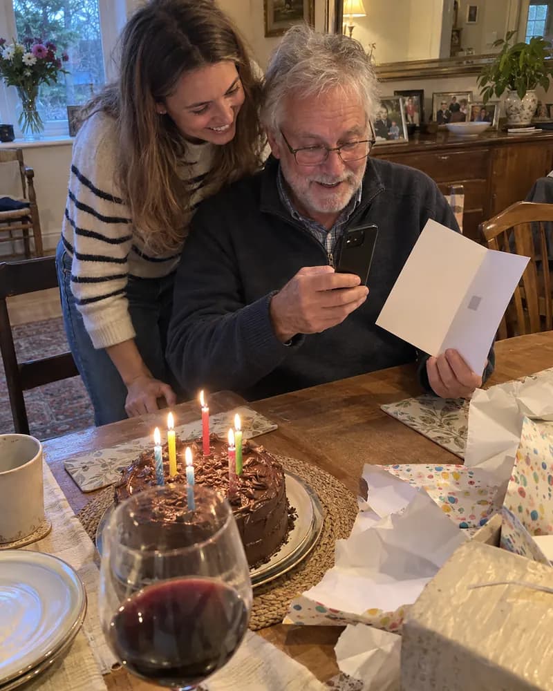 A man in his sixties at a dining table pointing his phone at a greeting card while his daughter leans in grinning, a birthday cake beside them (AI photo, fictional people)