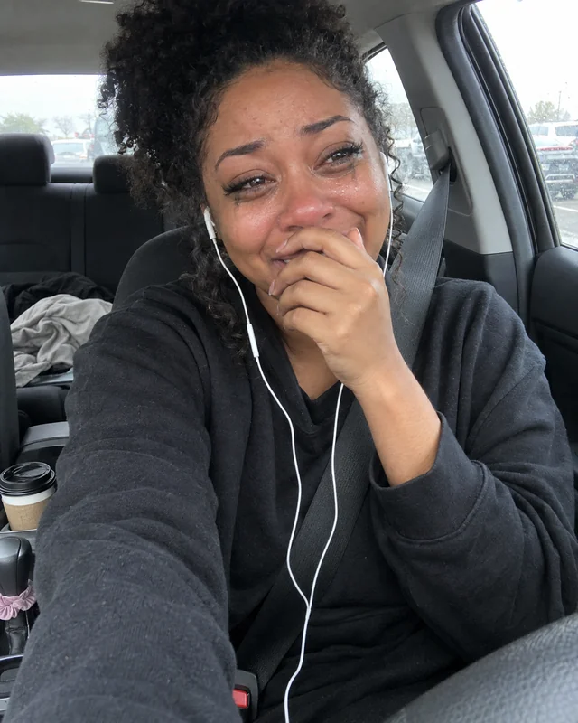 A young woman takes a teary selfie in her parked car with earbuds in, hand at her mouth, mascara smudged (AI photo, fictional person)