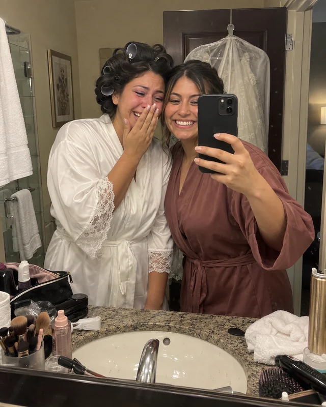 Two sisters take a mirror selfie on a wedding morning, the bride in a robe and hair rollers laughing through tears (AI photo, fictional people)