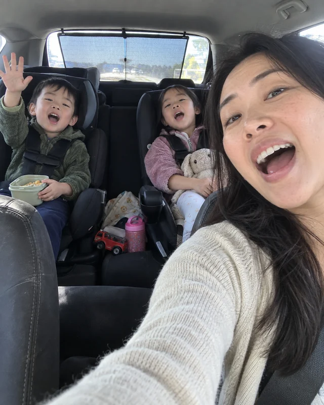 A mother takes a selfie in the car while her two small kids in car seats behind her sing along (AI photo, fictional people)