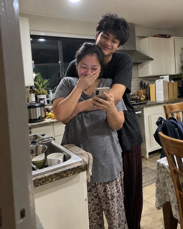 A mother in her kitchen covers her mouth while holding her phone as her teenage son hugs her from behind (AI photo, fictional people)