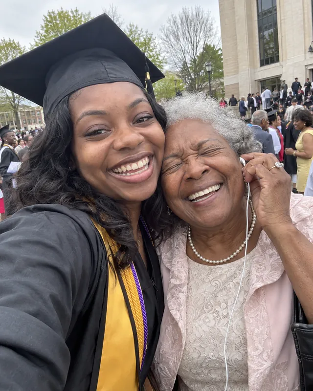 A graduate in cap and gown takes a selfie cheek to cheek with her laughing grandmother, who holds an earbud to her ear (AI photo, fictional people)