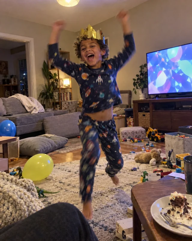 A young boy in pajamas and a paper crown jumps with his arms up in a messy living room (AI photo, fictional person)