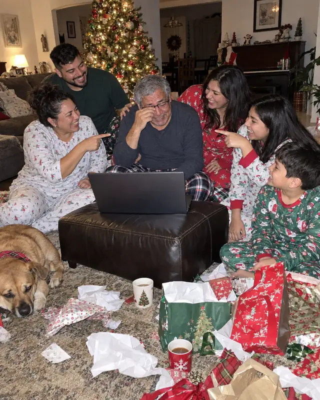 A family in holiday pajamas laughs and points at the grandfather wiping his eye in front of a laptop on Christmas morning (AI photo, fictional people)