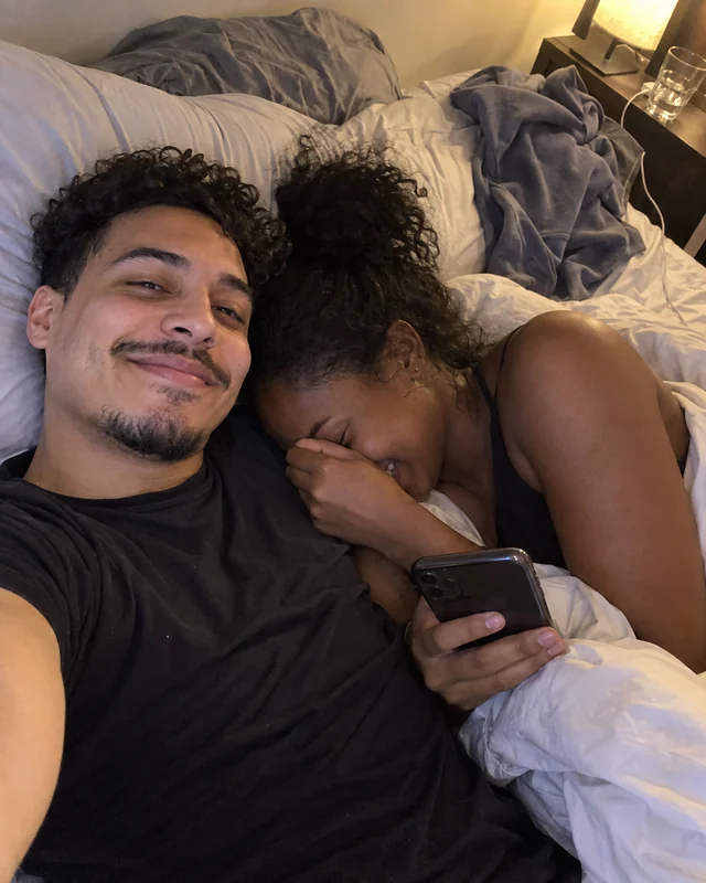 A man takes a selfie in bed while his girlfriend hides her laughing, teary face against his shoulder, holding his phone (AI photo, fictional people)