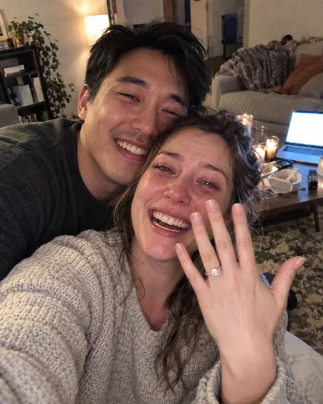 A woman with teary eyes shows her engagement ring in a selfie with her smiling fiance in their living room (AI photo, fictional people)