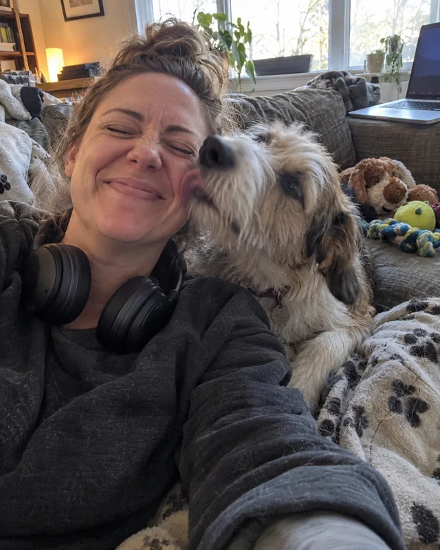 A woman with headphones around her neck laughs with her eyes shut as her scruffy dog licks her cheek on the couch (AI photo, fictional person)
