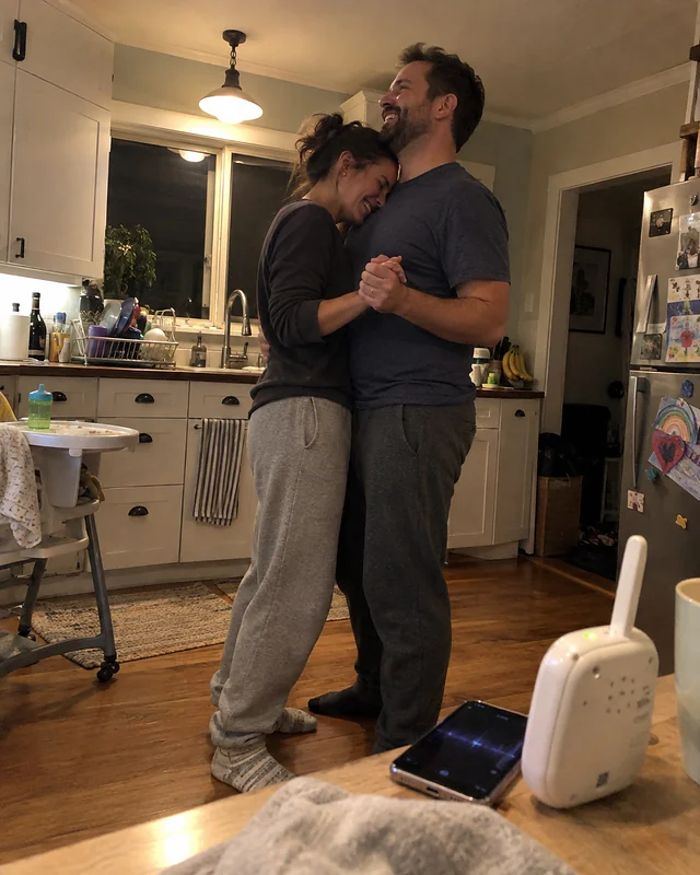 A couple in socks and sweatpants slow dancing in a messy kitchen at night, her forehead against his chest (AI photo, fictional people)