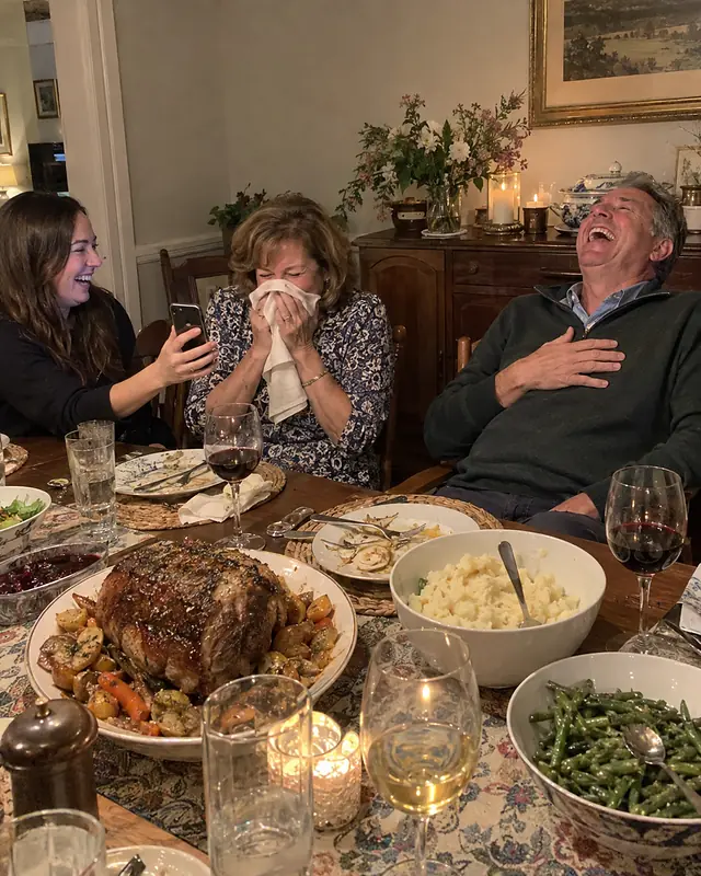 An older couple at a dinner table laughing hard while their adult daughter holds a phone toward them (AI photo, fictional people)