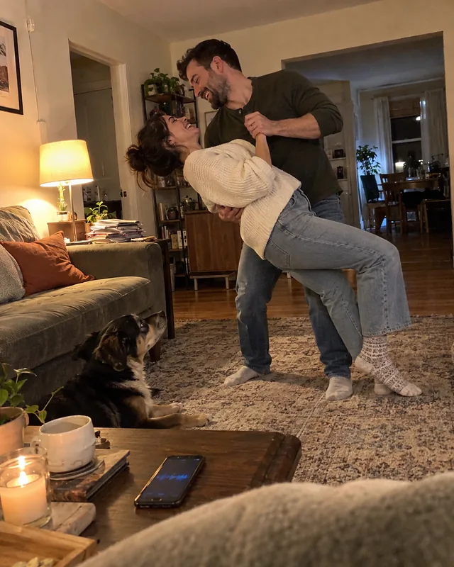 A couple dancing in socks in a small living room, he dips her and both laugh, a dog watching from the rug (AI photo, fictional people)