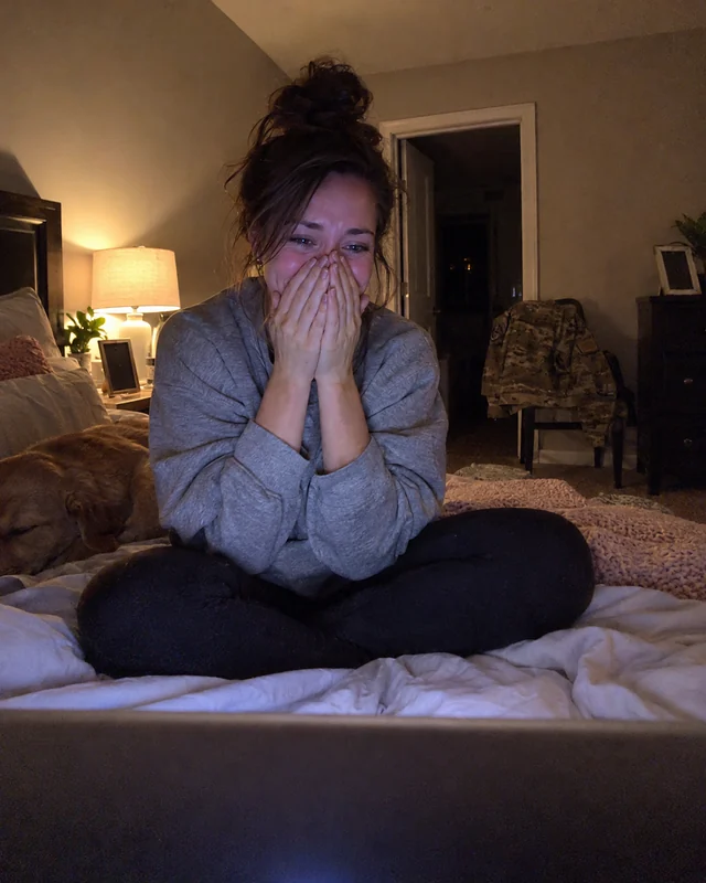 A woman sitting cross-legged on her bed at night in front of a glowing laptop, both hands over her mouth, laughing through tears (AI photo, fictional person)