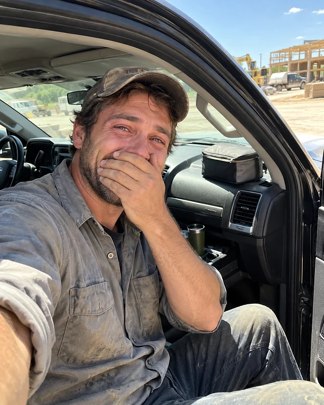 A man in a work shirt sits in his pickup truck at a job site with a hand over his mouth, eyes wet, grinning (AI photo, fictional person)