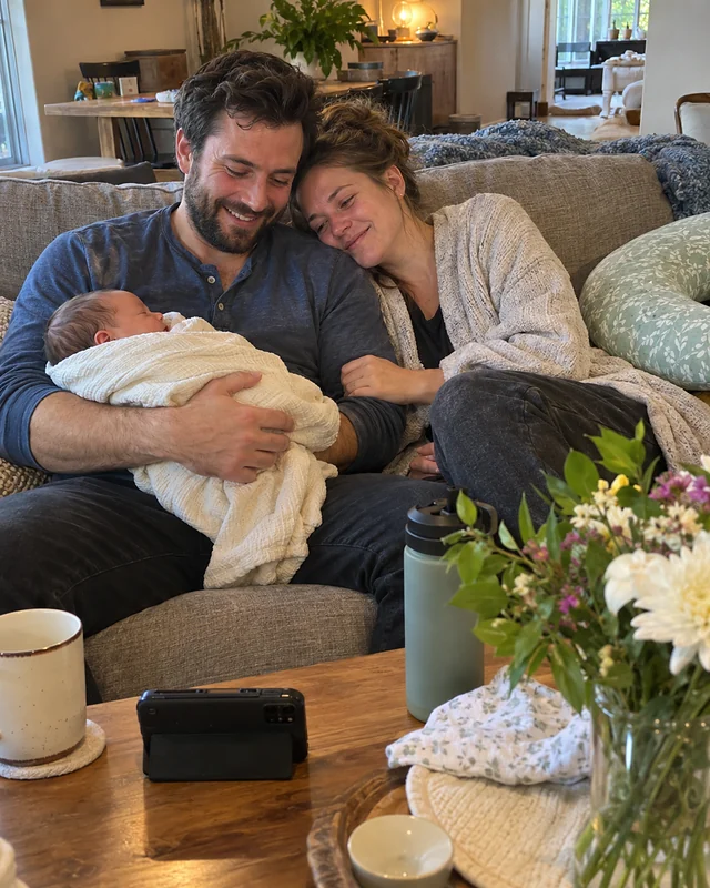 A new father on a sofa holds a sleeping newborn while the mother rests her head on his shoulder (AI photo, fictional people)