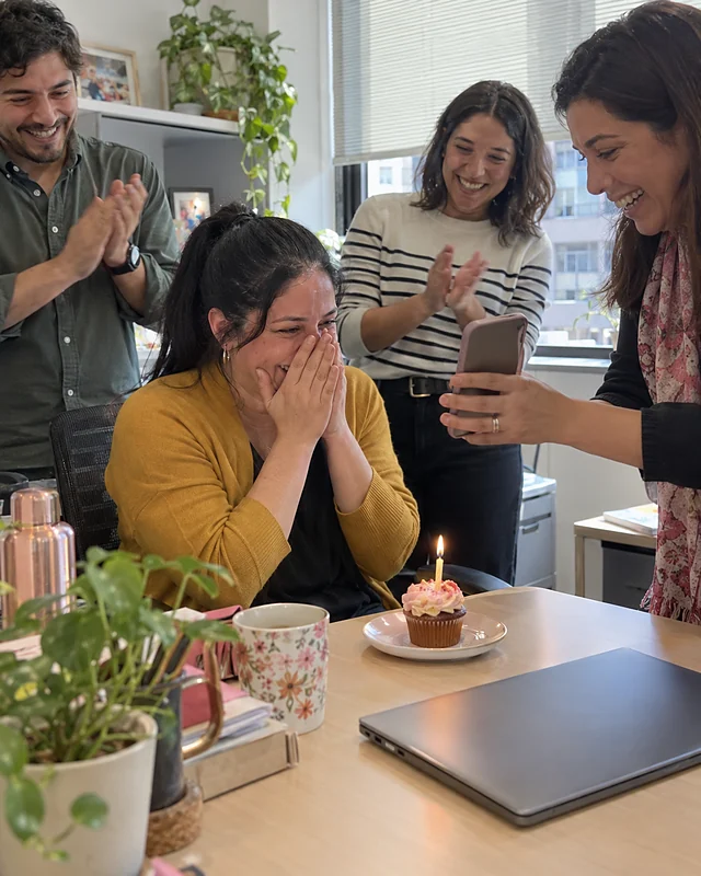 In a bright office, a woman in a mustard cardigan covers her face laughing in front of a cupcake with a candle, while three coworkers clap and one shows her a phone (AI photo, fictional people)