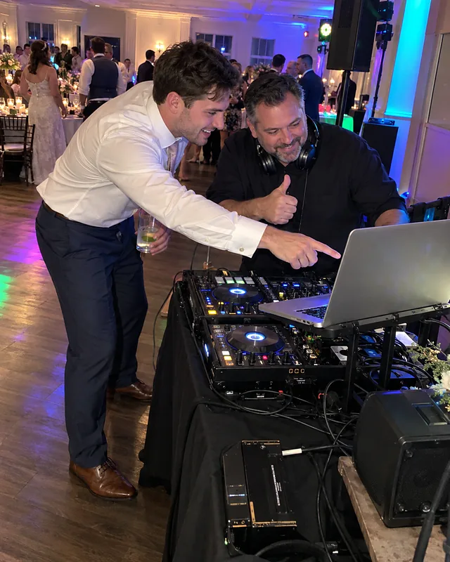 A groom in shirtsleeves points at a DJ's laptop at a reception while the DJ gives a thumbs up (AI photo, fictional people)