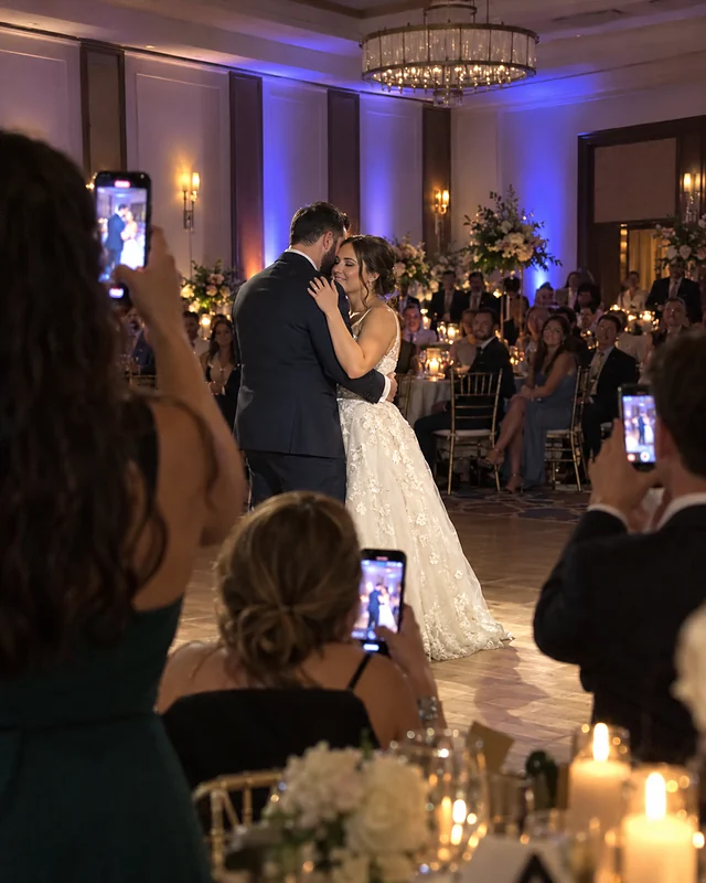 A guest's phone photo of a bride and groom slow dancing, seen past raised phones and shoulders (AI photo, fictional people)