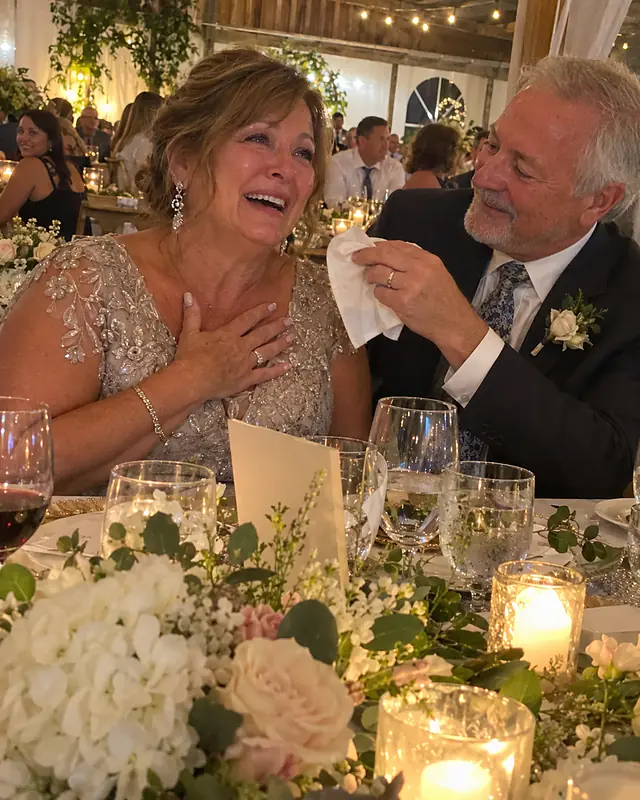 A woman in her late fifties at a reception table with her hand on her heart, crying and laughing as her husband hands her a napkin (AI photo, fictional people)