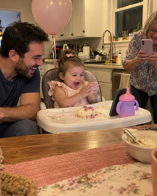 A one-year-old girl in a high chair claps and squeals at a phone playing music while her father laughs beside her (AI photo, fictional people)