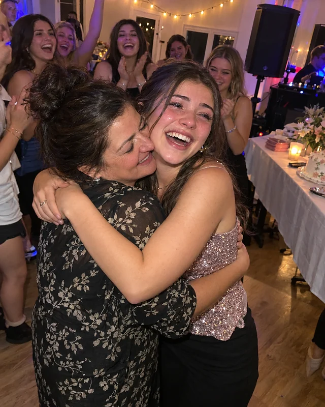 A sixteen-year-old girl hugs her mother tightly at her sweet sixteen, laughing with wet eyes, while friends cheer behind them (AI photo, fictional people)