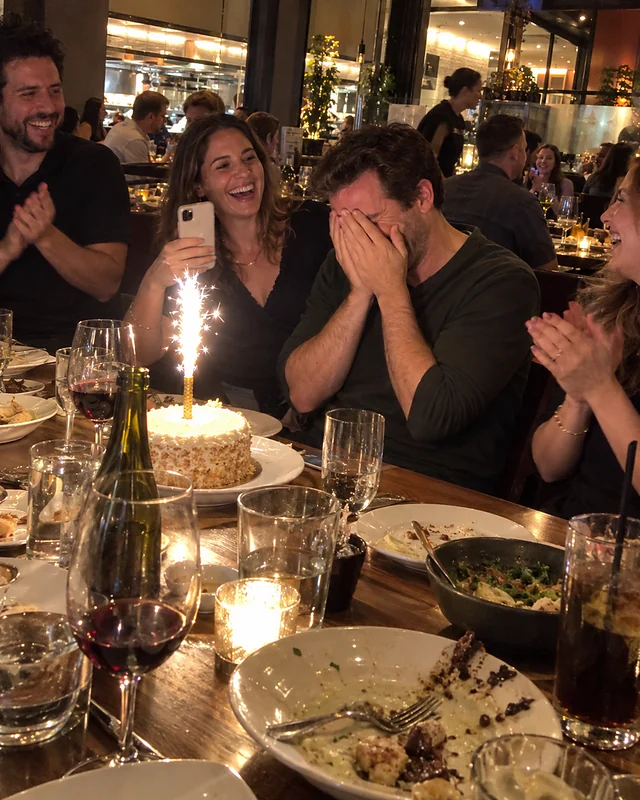 A man turning forty covers his face and laughs at a restaurant table while friends cheer and his wife holds up a phone (AI photo, fictional people)