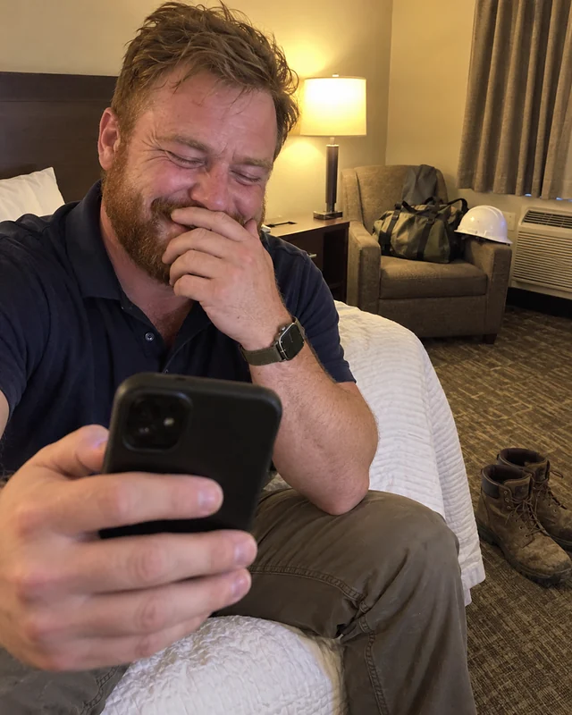 A man in a work polo sits in a motel room holding his phone up, laughing behind his hand, work boots on the floor (AI photo, fictional person)