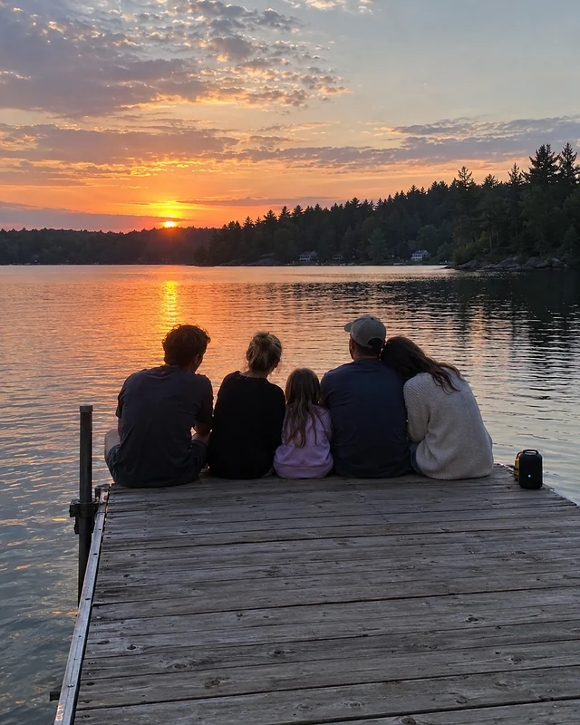 A family sits together at the end of a wooden lake dock at sunset, seen from behind, with a small speaker beside them (AI photo, fictional people)
