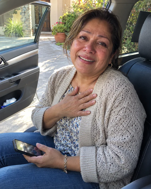 A woman turning sixty sits in a parked car with the door open, a hand on her chest and a phone in her lap, smiling with wet eyes (AI photo, fictional person)