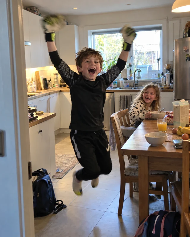 A nine-year-old boy in goalkeeper gloves jumps with both arms up in the kitchen while his little sister laughs at the table (AI photo, fictional people)