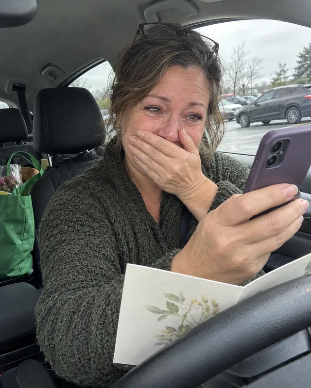 A woman in her fifties in a parked car holding a phone up to an opened greeting card, hand over her mouth (AI photo, fictional person)