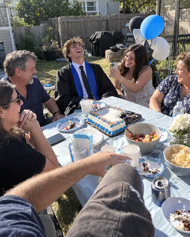 A young man in a graduation gown at a backyard table laughing at the phone his sister holds toward him (AI photo, fictional people)