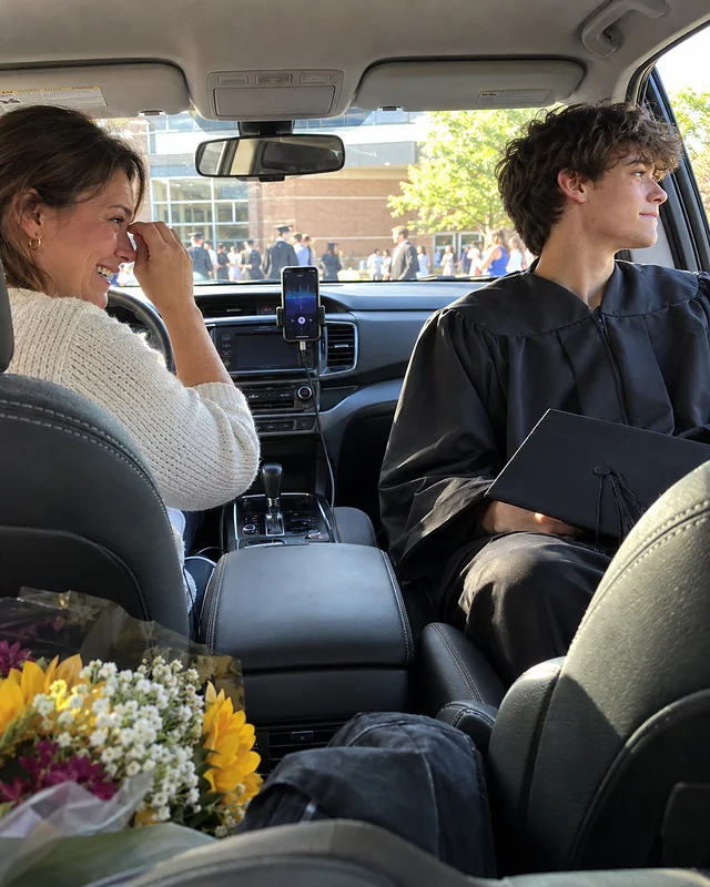 An eighteen-year-old in a graduation gown looks out the car window trying not to smile while his mother laughs and wipes her eye at the wheel (AI photo, fictional people)