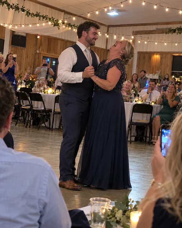 A groom grinning as he dances with his mother, who laughs with her head thrown back, guests holding up phones around them (AI photo, fictional people)