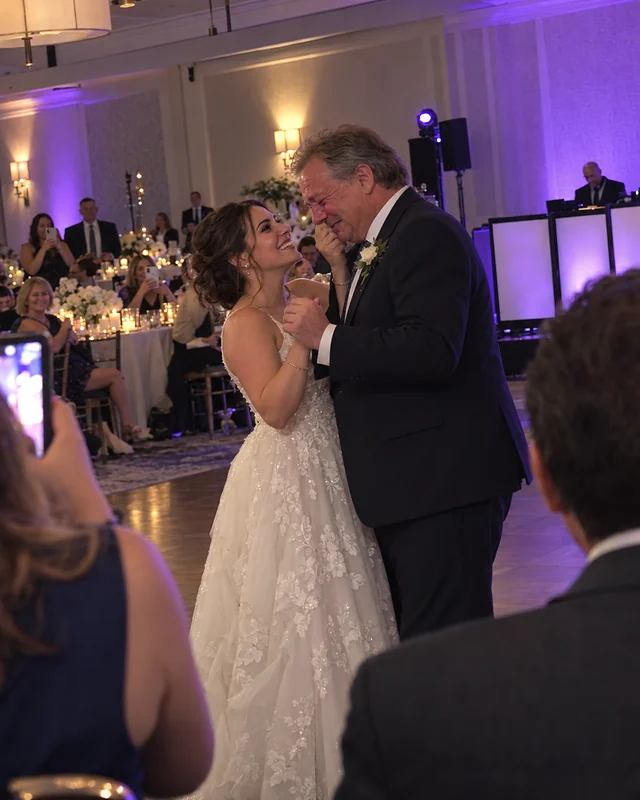 A father in a dark suit cries and laughs while dancing with his grinning daughter, the bride, as guests hold up phones at the edge of a ballroom (AI photo, fictional people)