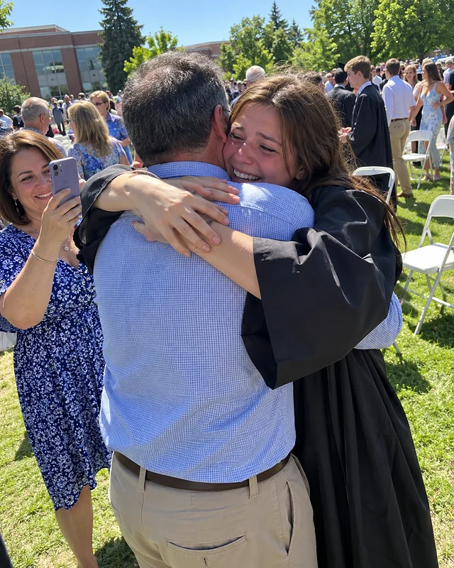 A graduate in a plain dark gown hugs her father tightly on a school lawn, crying and laughing, while her mother holds up a phone (AI photo, fictional people)