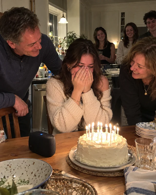 A young woman at a kitchen table covers her face, laughing, while her parents lean in beside a birthday cake with lit candles (AI photo, fictional people)