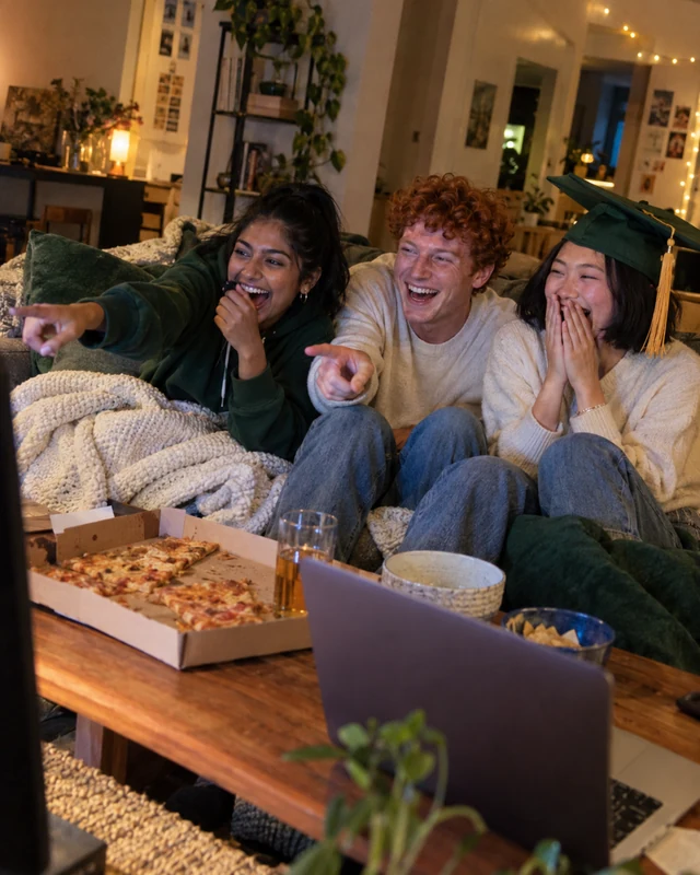 Three college roommates squeezed on a couch laugh and point at a glowing TV just out of frame, one wearing a green mortarboard with a gold tassel askew (AI photo, fictional people)